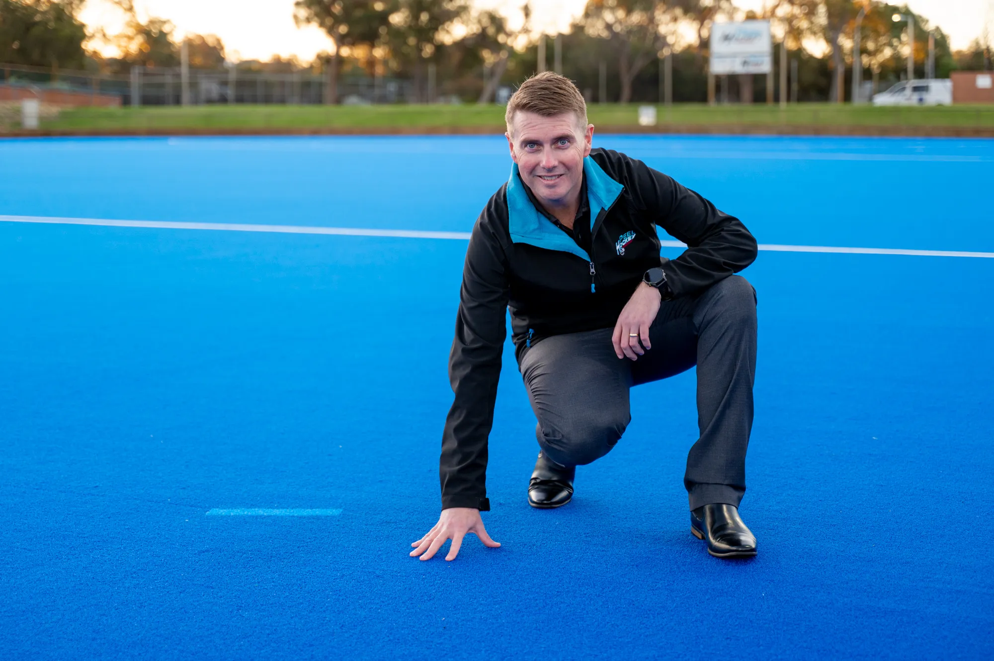 Person crouches on a bright blue hockey field, touching the turf with one hand. Wearing a black and blue jacket, grey trousers and black shoes, with trees, fencing and sports grounds blurred behind.